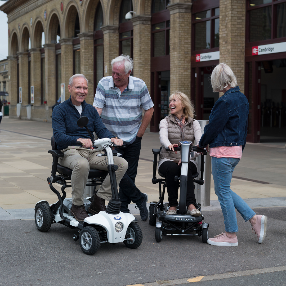 Showing the Maximo Plus Mobility Scooter being driven by a man with his wife driving another TGA scooter. The group are smiling and laughing as they drive through town.