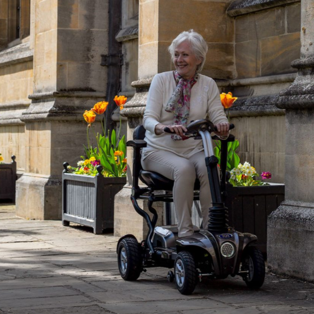 Showing a woman driving the TGA Maximo Mobility Scooter around town on the pavement, with a smile on her face.