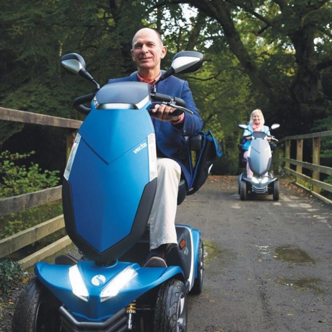 Showing the Rascal Vecta Sport being driven by a man in the park, with a woman riding behind him on her Vecta Sport also. Both people look happy and are comfortable riding on rough surfaces. 