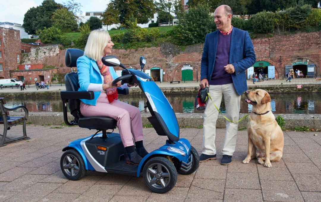 Showing the Rascal Vecta Sport Mobility Scooter being driven by a lady near a river, speaking to a man with his dog. They both are happy and engaged in a full conversation. 